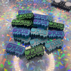 May include: A selection of small, rectangular PLUR charms in shades of teal, purple, and green. Each charm displays the word "PLUR" with a heart symbol. A miniature DJ mixer is visible in the background.