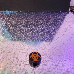 May include: A small, black, cylindrical container with an orange biohazard symbol on top. The container sits on a reflective surface with a star pattern. The background is dark.