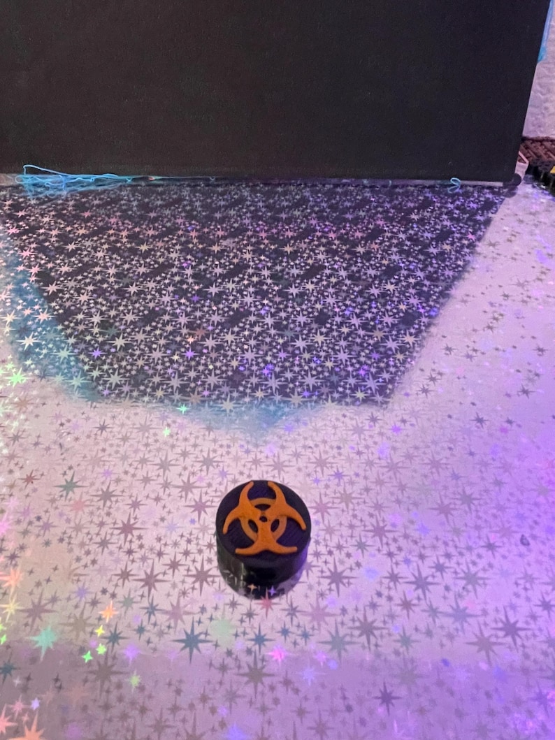 May include: A small, black, cylindrical container with an orange biohazard symbol on top. The container sits on a reflective surface with a star pattern. The background is dark.