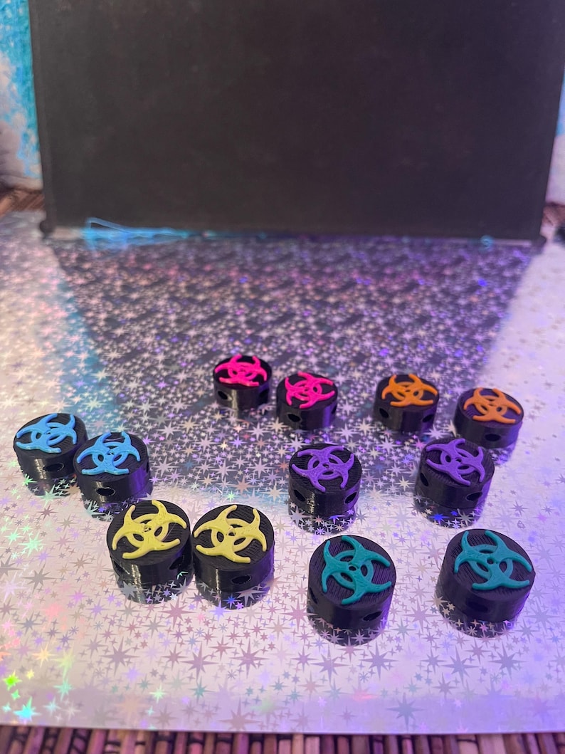 May include: A collection of black cylindrical objects, each topped with a colorful biohazard symbol in shades of blue, pink, orange, yellow, and teal. The objects are arranged on a shimmering, star-patterned surface.