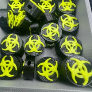 May include: A collection of black plugs with a neon yellow biohazard symbol on top. The plugs are cylindrical with a flat top and bottom. The biohazard symbol is a three-lobed design. The plugs are in a gray container.
