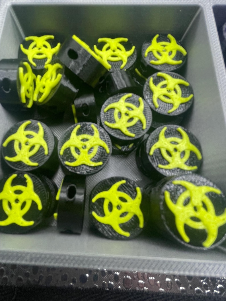 May include: A collection of black plugs with a neon yellow biohazard symbol on top. The plugs are cylindrical with a flat top and bottom. The biohazard symbol is a three-lobed design. The plugs are in a gray container.