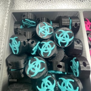 May include: A collection of black cylindrical objects with a teal biohazard symbol on top. The objects are arranged in a gray container, suggesting a product display or storage.