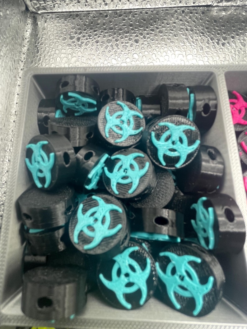 May include: A collection of black cylindrical objects with a teal biohazard symbol on top. The objects are arranged in a gray container, suggesting a product display or storage.