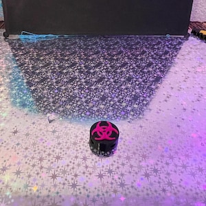 May include: A small, black container with a pink biohazard symbol on top. The container is cylindrical and sits on a reflective surface with a star pattern. The background is a dark, solid color.