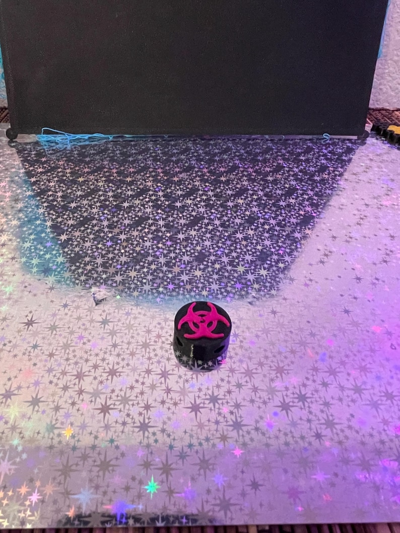 May include: A small, black container with a pink biohazard symbol on top. The container is cylindrical and sits on a reflective surface with a star pattern. The background is a dark, solid color.