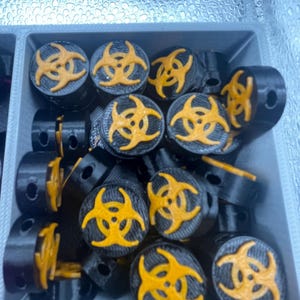 May include: A collection of black cylindrical objects with a yellow biohazard symbol on top. The objects are arranged in a gray container. The biohazard symbol is a bright yellow color.