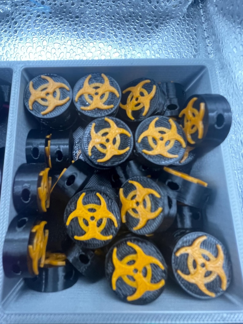 May include: A collection of black cylindrical objects with a yellow biohazard symbol on top. The objects are arranged in a gray container. The biohazard symbol is a bright yellow color.