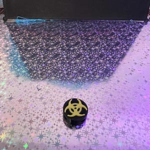 May include: A small, black, cylindrical object with a yellow biohazard symbol on top. The object is set against a reflective, star-patterned surface with a black backdrop.