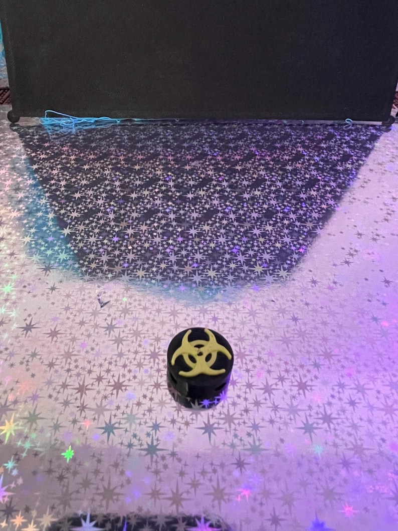 May include: A small, black, cylindrical object with a yellow biohazard symbol on top. The object is set against a reflective, star-patterned surface with a black backdrop.