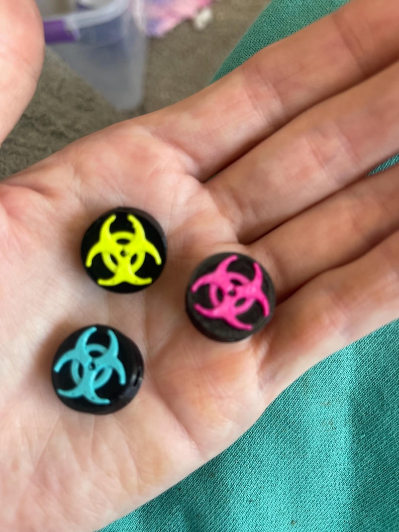 May include: Three black round buttons with a biohazard symbol in neon yellow, hot pink, and teal. The buttons are small and appear to be made of a glossy material. The biohazard symbols are centered on each button.