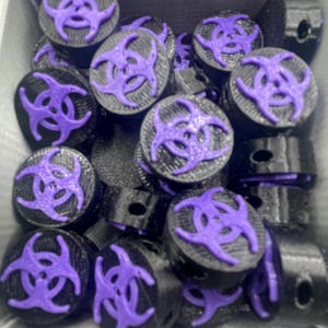 May include: A collection of black and purple decorative plugs with a biohazard symbol. The plugs are cylindrical with a flat top featuring a raised purple biohazard design. The items are in a gray container.