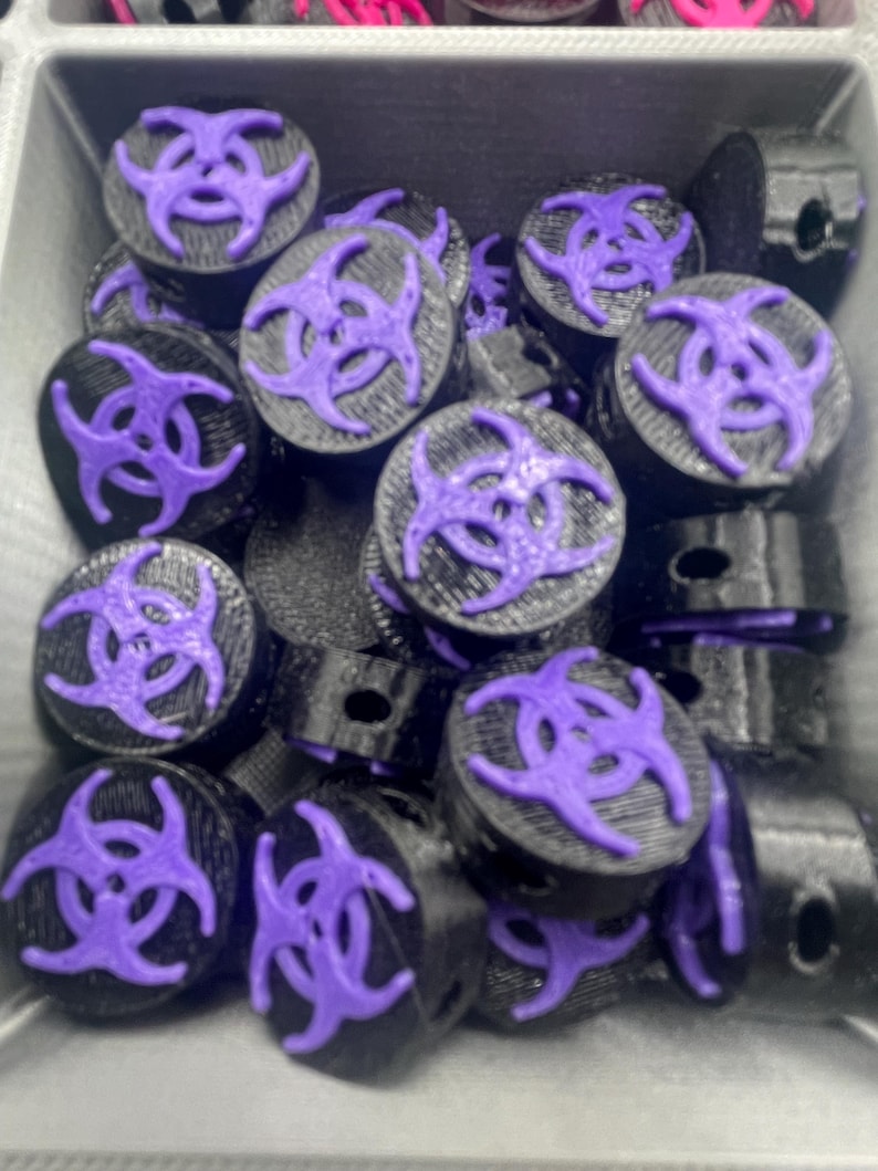 May include: A collection of black and purple decorative plugs with a biohazard symbol. The plugs are cylindrical with a flat top featuring a raised purple biohazard design. The items are in a gray container.