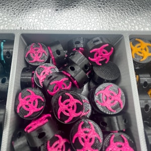 May include: A collection of black and pink biohazard symbol beads. The beads are cylindrical with a flat top featuring a bright pink biohazard symbol. The beads are contained in a gray plastic tray.