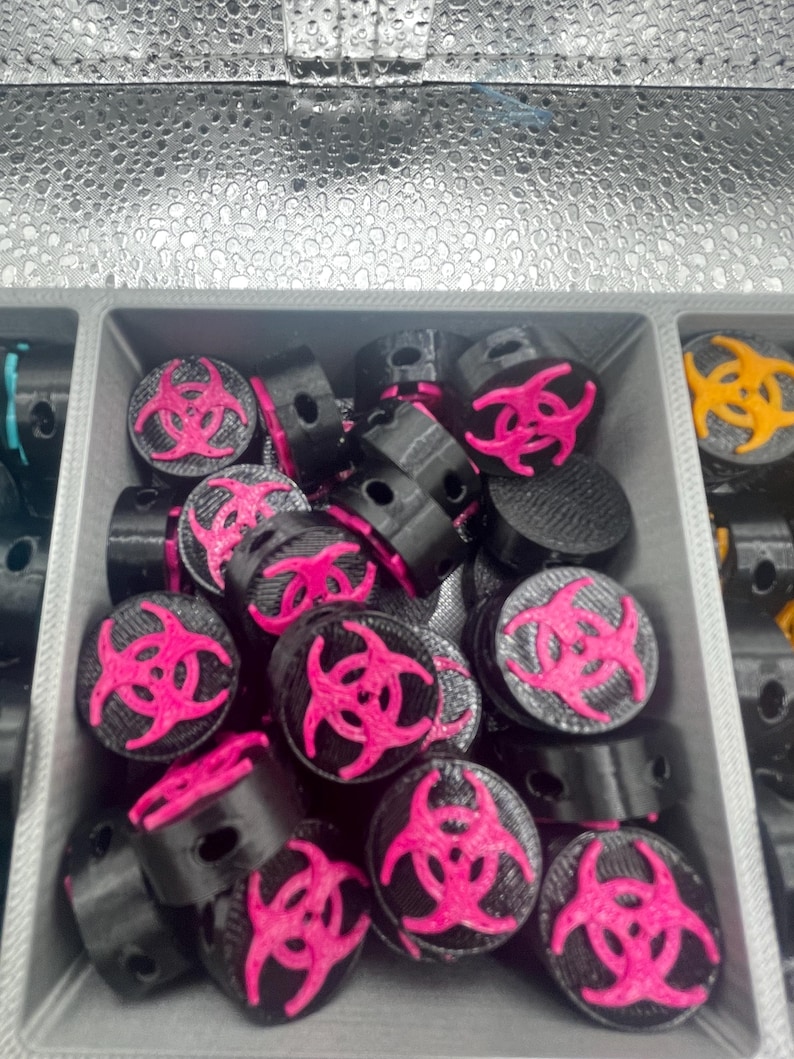 May include: A collection of black and pink biohazard symbol beads. The beads are cylindrical with a flat top featuring a bright pink biohazard symbol. The beads are contained in a gray plastic tray.