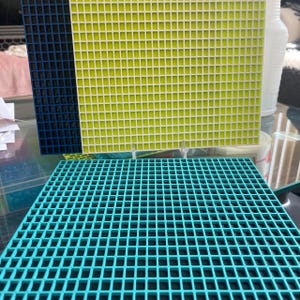 May include: A set of square grid mats featuring a teal mat in the foreground and a lime green mat positioned behind it. Each mat is constructed with a uniform pattern of small, open square cutouts arranged in a consistent, repeating grid layout.