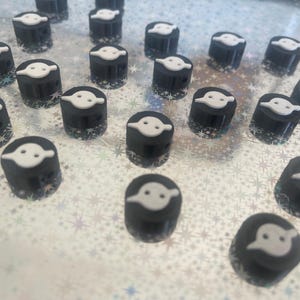 May include: A collection of small, cylindrical black objects with a white, stylized ghost design on top. The objects are arranged on a silver, star-patterned surface, creating a playful and thematic arrangement.