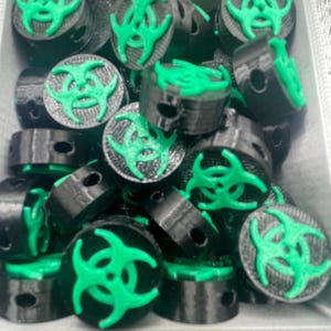 May include: A collection of black and green 3D-printed objects, each featuring a green biohazard symbol on top. The objects are cylindrical with a small hole on the side. They are arranged in a gray container.