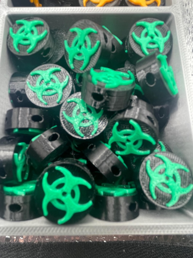 May include: A collection of black and green 3D-printed objects, each featuring a green biohazard symbol on top. The objects are cylindrical with a small hole on the side. They are arranged in a gray container.