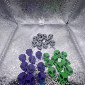 May include: A collection of alien-shaped objects in gray, purple, and green. Each alien has large, oval eyes and a smiling mouth. The objects are arranged on a reflective silver surface, creating a playful and whimsical aesthetic.