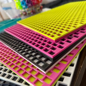 May include: A stack of rectangular plastic grid sheets in various colors including neon yellow, hot pink, black, and white. Each sheet features a uniform pattern of square cutouts arranged in a grid, with the edges showing a layered construction.