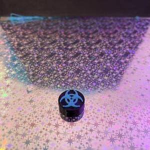 May include: A small, black container with a light blue biohazard symbol on top. The container is cylindrical and sits on a reflective surface with a star pattern. The background is dark.
