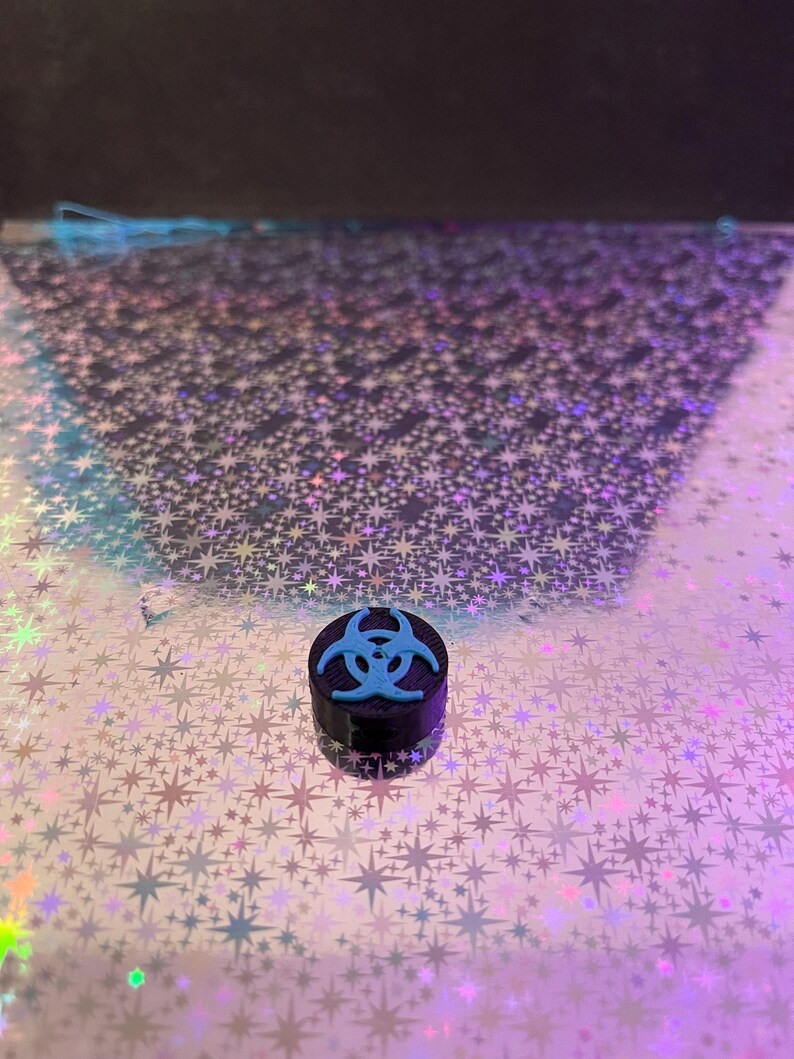 May include: A small, black container with a light blue biohazard symbol on top. The container is cylindrical and sits on a reflective surface with a star pattern. The background is dark.