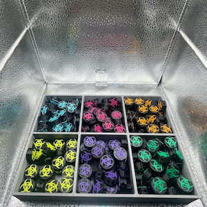 Pode incluir: Uma coleção de objetos cilíndricos pretos com um símbolo de perigo biológico em várias cores, incluindo azul, rosa, amarelo, roxo, laranja e verde. Os objetos estão dispostos numa bandeja cinza dividida, dentro de uma caixa prateada refletora.