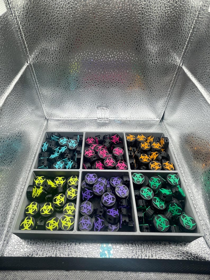 May include: A collection of black, cylindrical objects with a biohazard symbol in various colors, including blue, pink, yellow, purple, orange, and green. The objects are arranged in a gray divided tray, inside a silver reflective box.