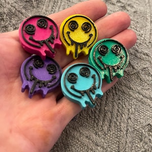 Puede incluir: Una mano sostiene seis coloridos pines redondos con caras sonrientes. Cada pin tiene un contorno negro y un diseño goteante. Los pines son en tonos rosa, amarillo, verde, morado, azul y turquesa.