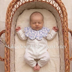 May include: A sleeping baby wearing a white onesie and a blue floral bib, lying in a wicker bassinet. The text "Makes Your Baby Even Cuter" is visible in the image.