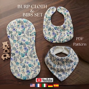 Puede incluir: Un conjunto de paño de eructos y dos baberos con estampado floral en tonos azul, morado y verde sobre fondo crema. El set incluye un paño largo, un babero redondeado, un babero tipo bandana triangular y tres pequeñas figuras de animales de madera.