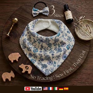 Op de afbeelding: Een driehoekig slabbetje met een blauw-wit bloemenpatroon en een witte fleece achterkant. De afbeelding bevat een bijpassende haarstrik, een klein flesje, een gouden rammelaar en drie houten dierfiguren met de tekst 3 Sizes.