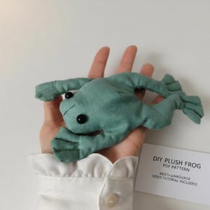 Può includere: Un peluche a forma di rana in tessuto verde salvia con occhi a perla neri, accompagnato da un cartoncino rettangolare bianco con il testo: DIY PLUSH FROG PDF PATTERN MULTI-LANGUAGE VIDEO TUTORIAL INCLUDED. La rana ha quattro arti palmati.