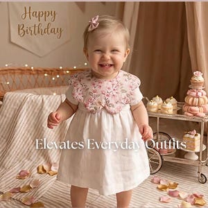 May include: A smiling child wearing a white dress with a floral collar and a matching bow. The image includes text that reads "Happy Birthday" and "Elevates Everyday Outfits". A decorative cart with desserts is in the background.