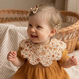 May include: A scalloped floral bib featuring a pattern of small orange, brown, and cream flowers on a light background, worn over a solid mustard yellow dress. A matching small floral hair bow is attached to the side of the head.