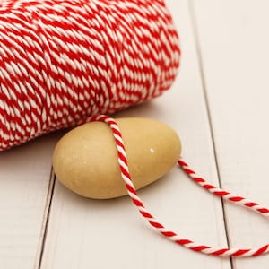 Op de afbeelding: Een rol rood-wit bakkersgaren naast een gladde, bruine steen. Het garen is om de steen gewikkeld, waardoor een lus ontstaat. De achtergrond is een wit houten oppervlak.
