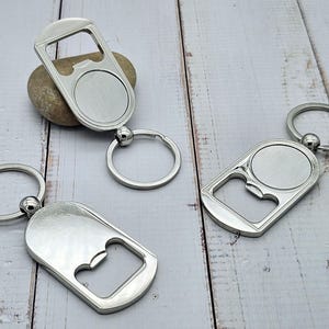 Peut inclure: Trois porte-clés décapsuleurs en métal argenté. Chaque porte-clés a un décapsuleur à une extrémité et une zone circulaire pour la personnalisation à l'autre. Les porte-clés sont sur une surface en bois clair.