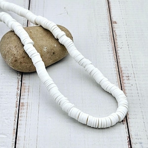Peut inclure: Un collier de perles blanches en forme de disques est posé sur une pierre lisse, brun clair. Le collier est composé de nombreuses petites perles plates et circulaires. Le fond est une surface en bois blanc.