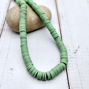 Peut inclure: Un collier composé de perles rondes et plates de couleur vert clair. Les perles sont enfilées pour former un collier. Le collier repose sur une pierre brun clair, posée sur une surface en bois blanc.