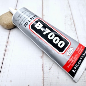 Peut inclure: Tube de colle multi-usages B-7000, argenté avec des détails noirs et rouges. Le tube affiche le texte "B-7000" et "110ml". La colle est imperméable, flexible et anti-vibration.