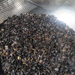 Op de afbeelding: Close-up van een metalen vergiet gevuld met donkere, geroosterde zonnebloempitten. De zaden zijn diep bruin van kleur en hebben een licht langwerpige vorm. Het vergiet heeft kleine, gelijkmatig verdeelde gaten.