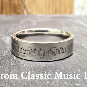 Op de afbeelding: Een zilverkleurige ring met gegraveerde muzieknoten en tekst. De ring heeft een band met muzieknotatie rondom de omtrek. De tekst "Custom Classic Music Band" staat onderaan de afbeelding.