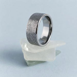 Könnte beinhalten: Ein metallischer Ring mit dunkelgrauem Finish, der ein komplexes, lasergraviertes Leiterplattenmuster auf der gesamten Außenfläche aufweist. Das Band hat eine glatte, polierte Innenseite und ein flaches Profil, das ein detailliertes elektronisches Design zeigt.