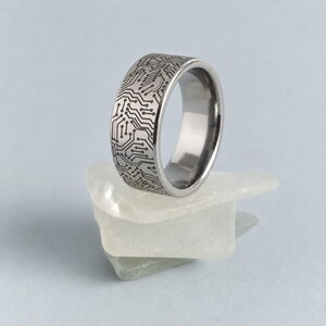 May include: A silver-toned metal ring featuring a dark, intricate circuit board pattern engraved across its entire outer surface. The band has a smooth, polished interior and a flat profile, showcasing a detailed technological design.