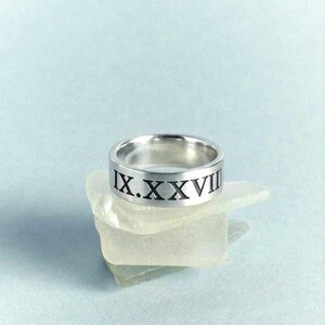Puede incluir: Un anillo de metal plateado con los números romanos IX.XXVII grabados en la banda exterior. El anillo tiene un acabado liso y pulido con un perfil plano, mostrando letras oscuras y claras que recorren toda la circunferencia del objeto.