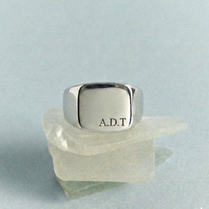 Puede incluir: Un anillo de sello plateado pulido con una cara cuadrada lisa y plana. La superficie del anillo está grabada con las letras mayúsculas A.D.T. en una fuente serif limpia, situadas hacia la esquina inferior derecha de la cara cuadrada.