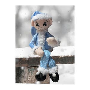 May include: A plush elf doll with a blue outfit and white trim, sitting on a snowy surface. The doll has a smiling face, a pointed hat, and striped blue and white legs. The elf is wearing black shoes with white trim.
