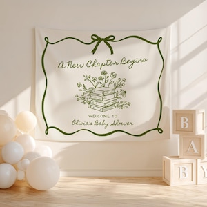 May include: A cream-colored baby shower banner with green text and a green border. The banner reads "A New Chapter Begins" and "Welcome to Olivia's Baby Shower." A stack of books with floral accents is in the center. Wooden blocks spell out "BABY."