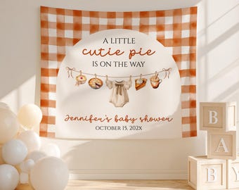 May include: A baby shower backdrop with a gingham pattern in shades of orange and white. The backdrop features the text "A LITTLE cutie pie IS ON THE WAY" and "Jennifer's baby shower OCTOBER 15, 202X". Decorative elements include baby clothes and food.